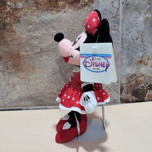 K&ouml;nnte beinhalten: Ein Minnie Mouse Pl&uuml;schtier von The Disney Store. Das Pl&uuml;schtier hat ein rotes Kleid mit wei&szlig;en Herzakzenten, wei&szlig;e Handschuhe und rote Schuhe. Die Schleife auf ihrem Kopf ist rot mit wei&szlig;en Herzen. Ein Etikett von The Disney Store ist angebracht.