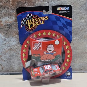 Könnte beinhalten: Ein NASCAR Winners Circle Spielzeug-Rennwagen, mit einem rot-orangen Farbschema und der Nummer 20. Die Verpackung enthält das The Home Depot-Logo und eine Cartoonfigur. Offiziell lizenziert von NASCAR.