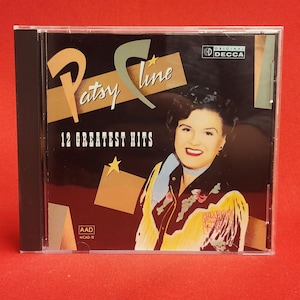 Op de afbeelding: Een cd-hoesje van Patsy Cline's "12 Greatest Hits". De hoes toont een portret van Patsy Cline, met de tekst "Patsy Cline" in een gestileerd lettertype. De cd-hoes heeft een zwarte rug en een doorzichtige plastic voorkant.