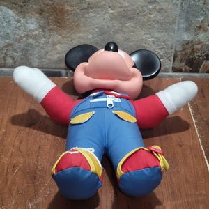 May include: A plush Mickey Mouse doll with a pink face, black ears, and a red shirt. The doll wears blue overalls with yellow accents and red and blue shoes. The arms are outstretched, and the doll is lying on a wooden surface.