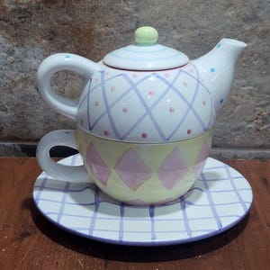 Puede incluir: Juego de té de cerámica decorativo que incluye tetera, taza y platillo. La tetera y la taza son blancas con un esquema de colores pastel, incluyendo patrones de diamantes y cuadrículas rosas, azules y amarillas. El platillo tiene un patrón de cuadrícula a juego.