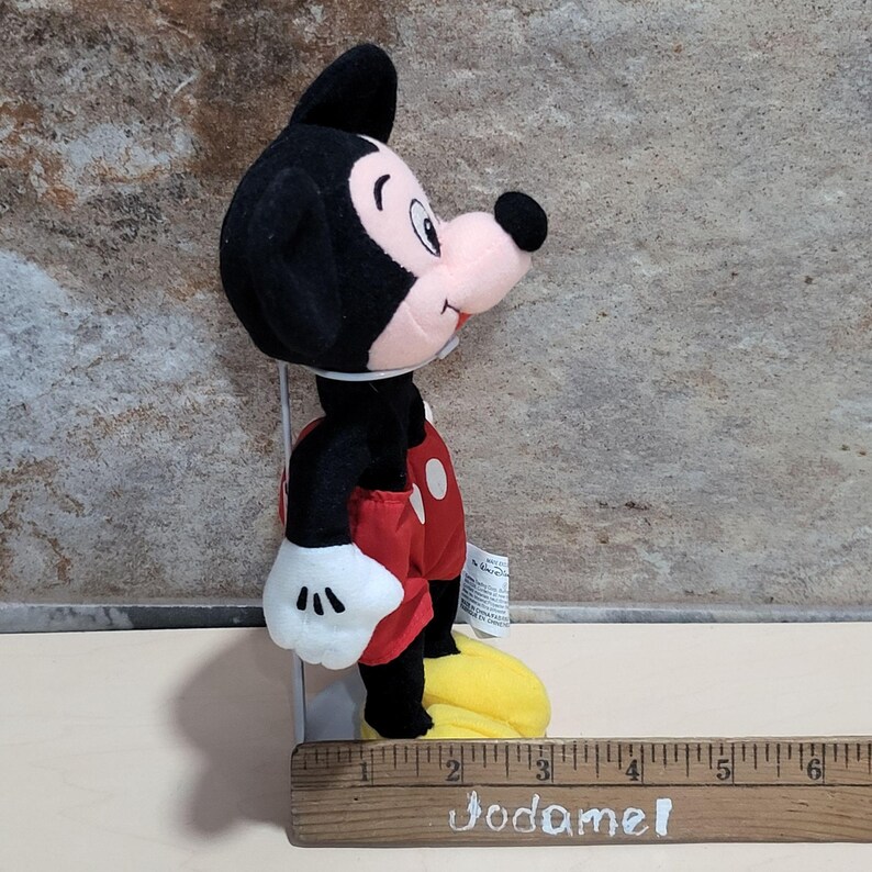 May include: A plush Mickey Mouse toy, featuring a black head and ears, a pink face, and a black nose. He wears red shorts with white buttons, white gloves, and yellow shoes. The toy is approximately 6 inches tall.