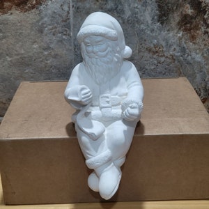 May include: White ceramic Santa Claus figurine sitting on a box. He is holding a scroll in one hand and a bell in the other.