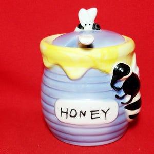 Puede incluir: Un tarro de miel de cerámica con un diseño de abeja y panal. El tarro es azul y amarillo con una etiqueta blanca que dice "HONEY".