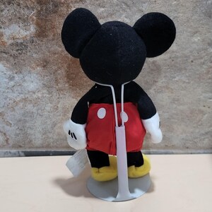 May include: A plush Mickey Mouse doll with black ears, a black head and arms, red shorts with white buttons, yellow shoes, and white gloves. The doll is standing on a gray stand.