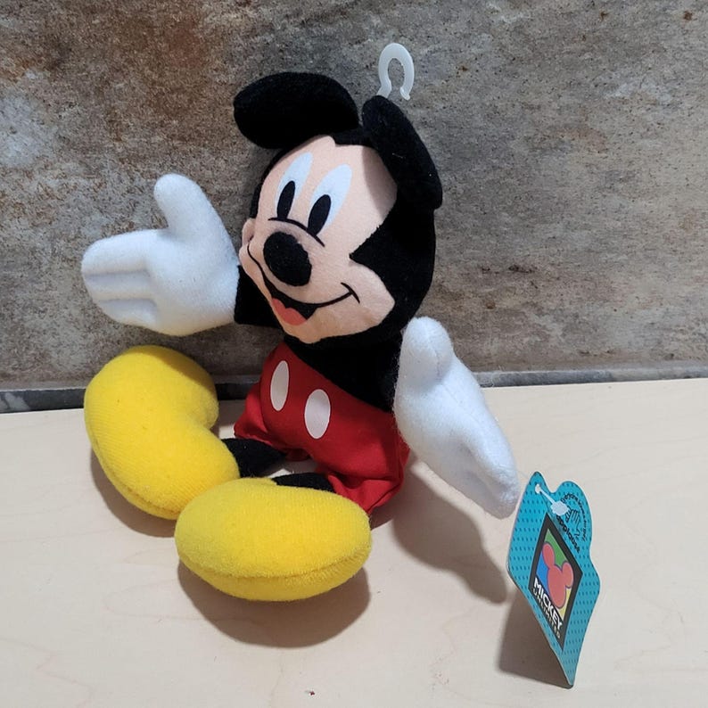 K&ouml;nnte beinhalten: Ein Pl&uuml;schtier von Mickey Mouse mit schwarzen Ohren, einem schwarzen Kopf und einem wei&szlig;en Gesicht. Das Spielzeug hat wei&szlig;e Handschuhe, rote Shorts mit wei&szlig;en Kn&ouml;pfen und gelbe Schuhe. Ein blaues Etikett mit dem Mickey Mouse-Logo ist angebracht.
