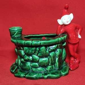 Könnte beinhalten: Ein grüner Keramik-Pflanztopf in Form eines Steinbrunnens mit einem kleinen Eimer an der Seite. Eine rote Elfenfigur lehnt sich an den Brunnen.