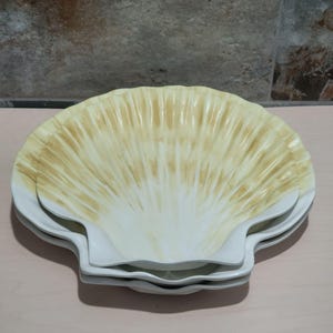 May include: A stack of three white scallop-shaped plates. The plates have a cream and white color scheme, with a radial pattern emanating from the center. The plates are stacked on top of each other.
