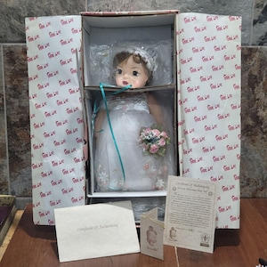 Peut inclure: Poupée Terri Lee dans une boîte, vêtue en mariée. La poupée porte une robe blanche avec des motifs floraux et un voile. Elle tient un bouquet de fleurs roses et blanches. La boîte a un motif floral et comprend un certificat d'authenticité.
