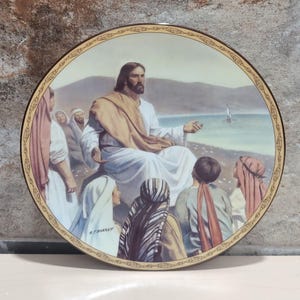 May include: A decorative plate featuring a religious scene. The image depicts a man in white robes and a tan shawl, seated and speaking to a group of people. The plate has a gold border with a decorative pattern.