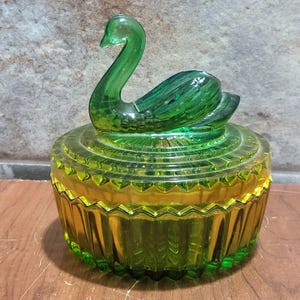 Könnte beinhalten: Eine grüne Glas-Schmuckdose mit einem Deckel in Form eines Schwans. Der Deckel ist strukturiert und facettiert, mit dem Kopf des Schwans zur Seite gedreht.