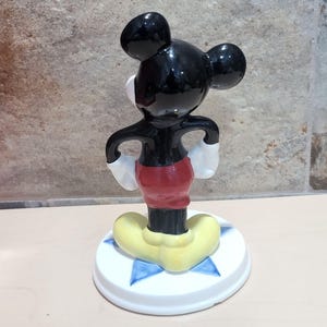 Puede incluir: Una figurita de cer&aacute;mica negra y blanca de Mickey Mouse de pie sobre una base blanca con estrellas azules. Mickey lleva pantalones cortos rojos y zapatos amarillos.
