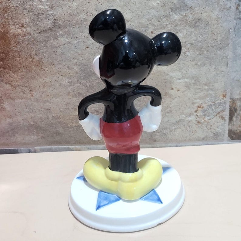 Puede incluir: Una figurita de cer&aacute;mica negra y blanca de Mickey Mouse de pie sobre una base blanca con estrellas azules. Mickey lleva pantalones cortos rojos y zapatos amarillos.