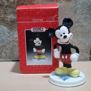 Puede incluir: Una figurita de cer&aacute;mica de Mickey Mouse de pie sobre una estrella azul. La figurita es negra con pantalones cortos rojos y zapatos amarillos. La caja dice "Schmid Disney Characters Figurine".