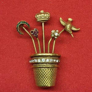 May include: A gold-toned thimble brooch with a crown, a heart, a crescent moon, a flower, and a hummingbird on top. The thimble is decorated with rhinestones.