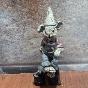 Pode incluir: Uma estatueta extravagante de um coelho usando um chapéu de burro com a palavra "DUNCE". O coelho está vestido com macacão azul e uma camisa listrada vermelha e branca. Um pequeno sapo está sentado ao lado de um balde na base da estatueta.