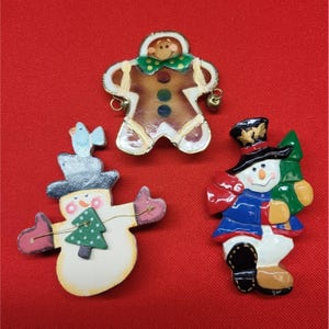 May include: Three Christmas-themed brooches on a red background. The brooches include a gingerbread man, a snowman with a bird, and a snowman holding a Christmas tree. The gingerbread man has a green bow tie and gold accents. The snowman brooches have painted details.