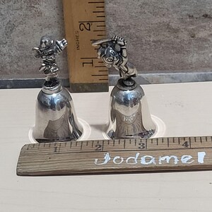 Pu&ograve; includere: Due campane color argento con figurine dettagliate sulla parte superiore. Una campana presenta un giocatore di baseball, l'altra un giocatore di football americano. Le campane sono alte circa 10 cm e poggiano su basi di plastica trasparente. Un righello di legno &egrave; in primo piano.