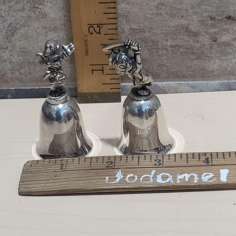 Pu&ograve; includere: Due campane color argento con figurine dettagliate sulla parte superiore. Una campana presenta un giocatore di baseball, l'altra un giocatore di football americano. Le campane sono alte circa 10 cm e poggiano su basi di plastica trasparente. Un righello di legno &egrave; in primo piano.