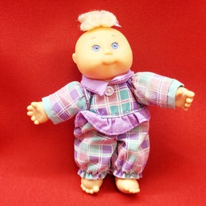May include: A vintage baby doll with a bald head, blue eyes, and a small tuft of blonde hair. The doll is wearing a plaid pajama set in shades of purple, teal, and light blue. The pajamas have a ruffled collar and cuffs.