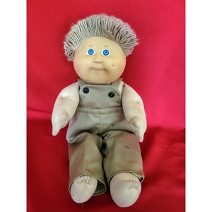 May include: A vintage Cabbage Patch Kid doll with blonde hair and blue eyes. The doll is wearing a brown overall outfit.