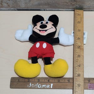 K&ouml;nnte beinhalten: Ein Pl&uuml;schtier von Mickey Mouse, mit schwarzem Kopf und Ohren, roter Hose, gelben Schuhen und wei&szlig;en Handschuhen. Das Spielzeug ist etwa 18 cm hoch und hat ein Etikett.