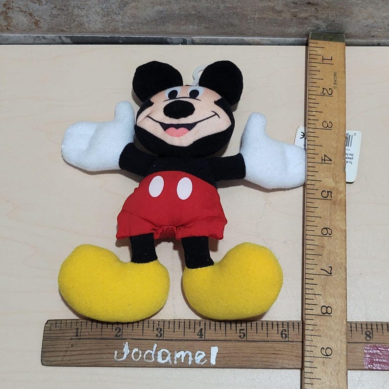 K&ouml;nnte beinhalten: Ein Pl&uuml;schtier von Mickey Mouse, mit schwarzem Kopf und Ohren, roter Hose, gelben Schuhen und wei&szlig;en Handschuhen. Das Spielzeug ist etwa 18 cm hoch und hat ein Etikett.