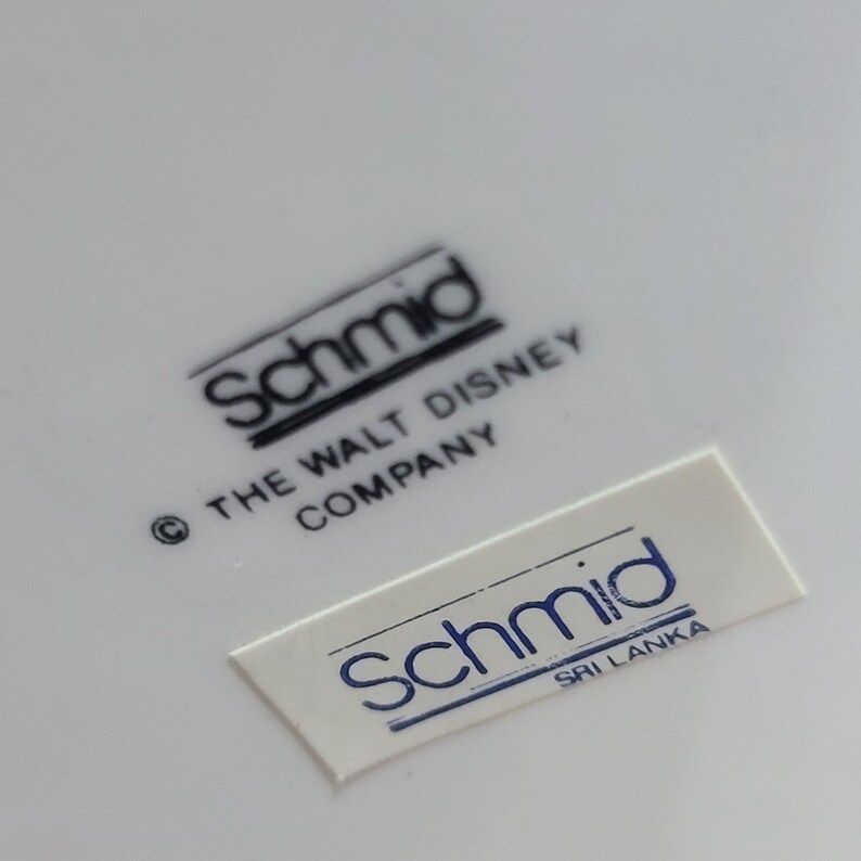 Puede incluir: Primer plano de un plato de cer&aacute;mica blanco con dos etiquetas en blanco y negro. La etiqueta superior dice "Schmid &copy; THE WALT DISNEY COMPANY". La etiqueta inferior dice "Schmid SRI LANKA".