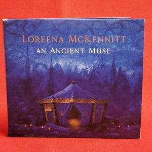Könnte beinhalten: Ein CD-Cover mit dem Album "An Ancient Muse" von Loreena McKennitt. Das Cover zeigt ein blaues Zelt mit goldenen Akzenten vor einer dunkelblauen, mystischen Landschaft. Der Name des Künstlers und der Albumtitel sind in Gold geschrieben.