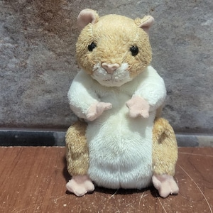 Peut inclure: Une peluche en forme de hamster avec un pelage doux et texturé dans des tons beige clair et blanc. Ce jouet en peluche présente de petites oreilles arrondies, des yeux en perles sombres, un nez rose pâle et de petites pattes tendues vers l'avant.
