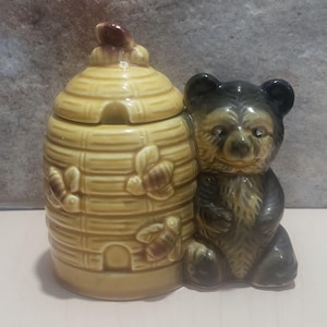 May include: A ceramic honey pot shaped like a beehive with a bear figurine. The beehive is yellow with brown bee and butterfly accents. The bear is dark brown and gray.
