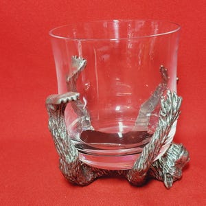May include: A clear glass tumbler with a silver monkey figurine base. The monkey is holding the glass with its arms and legs.