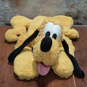 May include: A plush toy of the character Pluto, featuring yellow textured fur, long black ears, a large black nose, white eye patches with black pupils, and a pink tongue hanging out. The character is depicted in a lying down position.