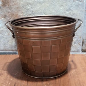May include: A copper-colored metal planter with two handles and a checkered pattern. The planter is round and has a slightly flared rim.
