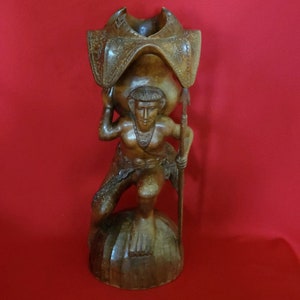 May include: A wooden sculpture of a person carrying a large bowl on their back. The sculpture is carved in a detailed style and has a dark brown colour.