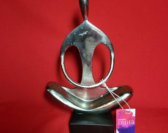 May include: A silver-colored metal sculpture of a person in a seated yoga pose. The sculpture has a rounded base and a tall, curved headpiece. It sits on a black rectangular base. A tag with the word "India" is attached.