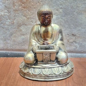 May include: A silver-colored Buddha statue sitting in a meditative pose, holding a small box with Chinese characters. The statue is on a base with intricate details.