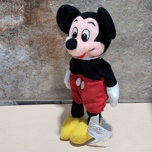 May include: A plush Mickey Mouse toy, featuring a black body, red shorts, and yellow shoes. The iconic character has a pink face, black ears, and white gloves. A white tag is attached to the toy's foot.