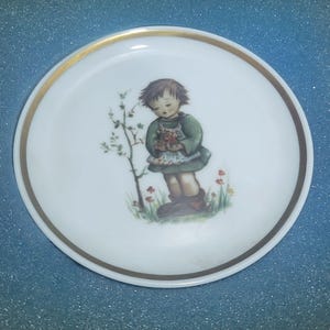 Può includere: Un piatto in porcellana bianca con un bordo dorato presenta un'immagine dipinta di una bambina con un vestito verde che tiene un mazzo di fiori. La bambina è in piedi in un campo di erba verde e fiori rossi.