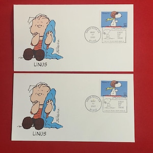 May include: Two white envelopes featuring Peanuts characters. One envelope shows Linus holding his blue blanket, with the name "LINUS" below. The other envelope has a Snoopy stamp and a "First Day of Issue" postmark from Santa Rosa, CA.