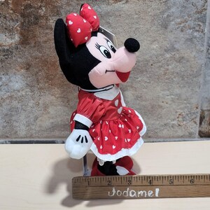 K&ouml;nnte beinhalten: Minnie Mouse Pl&uuml;schpuppe mit rotem Kleid mit wei&szlig;en Herzakzenten und passender Schleife. Die Puppe hat einen schwarzen Kopf und Ohren, ein rosa Gesicht und wei&szlig;e Handschuhe. Die Puppe steht auf einem Holzlineal.