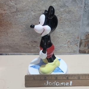 Puede incluir: Una figurita de cer&aacute;mica de Mickey Mouse de pie sobre una base azul en forma de estrella. Mickey lleva sus pantalones cortos rojos caracter&iacute;sticos y zapatos amarillos. La figurita mide aproximadamente 10 cm de alto.