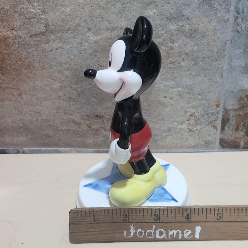 Puede incluir: Una figurita de cer&aacute;mica de Mickey Mouse de pie sobre una base azul en forma de estrella. Mickey lleva sus pantalones cortos rojos caracter&iacute;sticos y zapatos amarillos. La figurita mide aproximadamente 10 cm de alto.