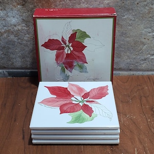 May include: Set of four white ceramic coasters featuring a red and green poinsettia design. The coasters are stacked and come in a red box with a poinsettia design.