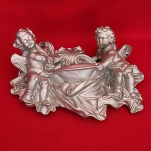 Könnte beinhalten: Eine silberfarbene Wanddekoration mit zwei Cherubinen, die eine Schale halten. Die Cherubinen sitzen auf einem drapierten Stoff mit verzierten Details.