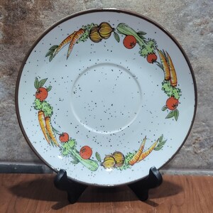 May include: White ceramic saucer with a brown rim and a black speckled glaze. The saucer has a ring of vegetables painted around the edge, including carrots, onions, tomatoes, and celery.