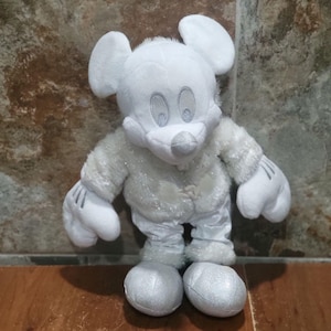Puede incluir: Un peluche de un personaje con orejas grandes y redondas, que viste una chaqueta blanca y difusa con detalles de purpurina plateada, pantalones blancos y zapatos blancos. El personaje tiene pelaje blanco, guantes blancos y una nariz plateada.
