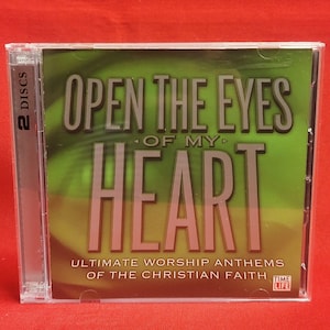 Puede incluir: Una caja de CD con el texto "OPEN THE EYES OF MY HEART" en letras grandes y en negrita. La caja también incluye el texto "ULTIMATE WORSHIP ANTHEMS OF THE CHRISTIAN FAITH" y el logotipo de Time Life. La caja es transparente con un fondo verde y amarillo.