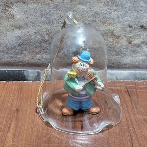 May include: A clear glass bell ornament with a gold string. Inside, a small clown figurine plays a violin. The clown wears a blue hat, green jacket, and blue shorts. The ornament has a scalloped edge.