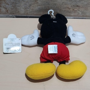 K&ouml;nnte beinhalten: Ein Pl&uuml;schtier von Mickey Mouse, von hinten gesehen, mit schwarzem Kopf und K&ouml;rper, roter Hose, wei&szlig;en Handschuhen und gelben Schuhen. Das Spielzeug hat ein Anh&auml;ngeetikett. Das Spielzeug wird von Applause hergestellt.